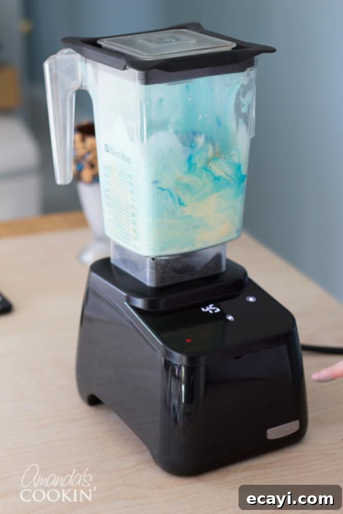 Cookie Monster Freakshakes are fun blue milkshakes topped with Cookie Monster cookies and surrounded by chocolate chip cookie pieces! #cookiemonster #cookiemonstershake #cookiemonsterparty #cookiemonsterrecipes #cookiemonsterfood #freakshake #cookies #icecream #milkshake #shakerecipes #sesamestreet #blueshakes  A blender with vanilla ice cream and milk, with a small amount of sky blue gel food coloring being added from a tube. This step highlights the process of tinting the milkshake base to achieve Cookie Monster's signature blue color.