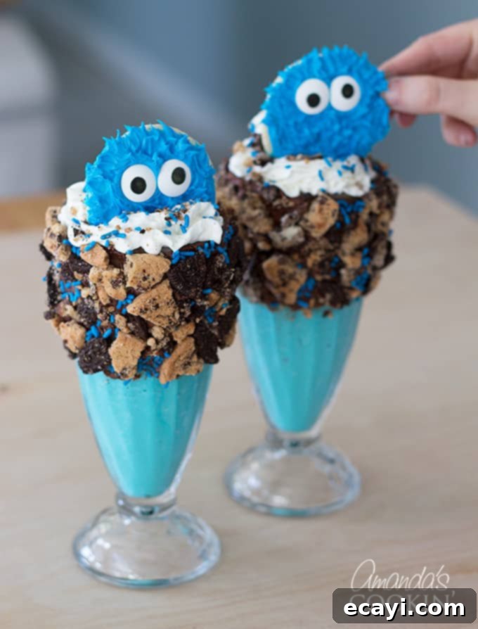 Cookie Monster Freakshakes are fun blue milkshakes topped with Cookie Monster cookies and surrounded by chocolate chip cookie pieces! #cookiemonster #cookiemonstershake #cookiemonsterparty #cookiemonsterrecipes #cookiemonsterfood #freakshake #cookies #icecream #milkshake #shakerecipes #sesamestreet #blueshakes  An adorable Cookie Monster sugar cookie with googly eyes being placed gently onto the whipped cream topping of a blue freak shake, surrounded by sprinkles and cookie crumbles. This image captures the final, signature step of assembling the themed dessert.