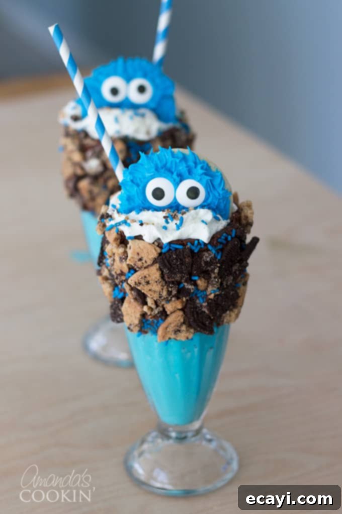 Cookie Monster Freakshakes are fun blue milkshakes topped with Cookie Monster cookies and surrounded by chocolate chip cookie pieces! #cookiemonster #cookiemonstershake #cookiemonsterparty #cookiemonsterrecipes #cookiemonsterfood #freakshake #cookies #icecream #milkshake #shakerecipes #sesamestreet #blueshakes  A close-up of a Cookie Monster Freak Shake, featuring a blue milkshake base, a generous swirl of whipped cream, blue sprinkles, and crushed chocolate chip cookies around the rim. A small, adorable Cookie Monster cookie with googly eyes sits on top, with more crushed cookies and a colorful striped straw, emphasizing its fun and festive nature.