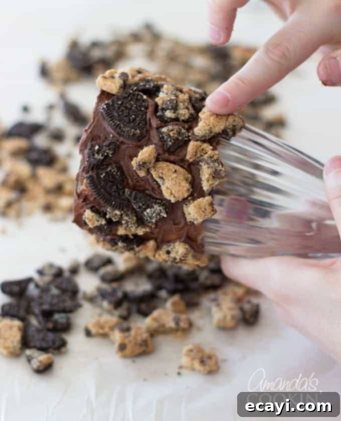 Cookie Monster Freakshakes are fun blue milkshakes topped with Cookie Monster cookies and surrounded by chocolate chip cookie pieces! #cookiemonster #cookiemonstershake #cookiemonsterparty #cookiemonsterrecipes #cookiemonsterfood #freakshake #cookies #icecream #milkshake #shakerecipes #sesamestreet #blueshakes  A hand gently rolling a chocolate-frosted milkshake glass into a pile of cookie crumbles spread on parchment paper, demonstrating the technique for coating the rim of the glass with cookie pieces. The image highlights the careful application required.