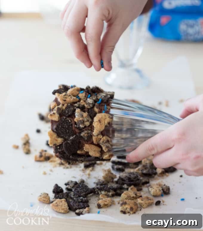 Cookie Monster Freakshakes are fun blue milkshakes topped with Cookie Monster cookies and surrounded by chocolate chip cookie pieces! #cookiemonster #cookiemonstershake #cookiemonsterparty #cookiemonsterrecipes #cookiemonsterfood #freakshake #cookies #icecream #milkshake #shakerecipes #sesamestreet #blueshakes  A hand carefully pressing blue sprinkles onto the chocolate-frosted and cookie-crusted rim of a milkshake glass, demonstrating the final decorative touch before chilling. The sprinkles fill in gaps between the cookie crumbles, adding color and sparkle.