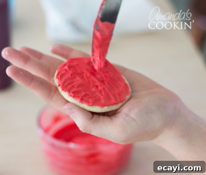 Elmo's Ultimate Freakshakes 4 Close-up of a hand spreading red vanilla frosting onto a sugar cookie, demonstrating the technique for Elmo's 'fur'.