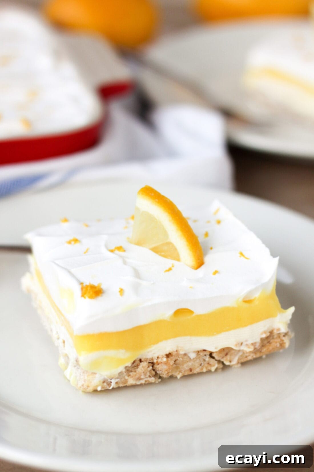 Lemon Lush. It's light and refreshing with 4 layers consisting of bottom pecan crust, lemon cream cheese, lemon pudding and topped with a whipped layer garnished with lemon zest. #lemonlush #lemondessert #lemonlasagna #nobake #onepandesserts #potluckdesserts #creamcheesedesserts #puddingdesserts  Piece of Lemon Lush on a plate