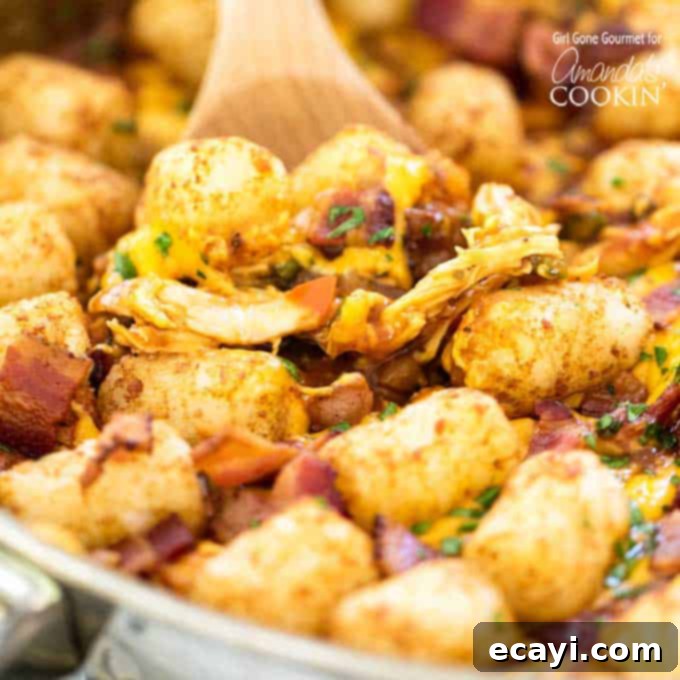 A close-up of the hearty BBQ Chicken Tater Tot Skillet ready to be served. BBQ chicken, tater tots, and crispy bacon piled high in a cast iron skillet with a wooden spoon inserted.