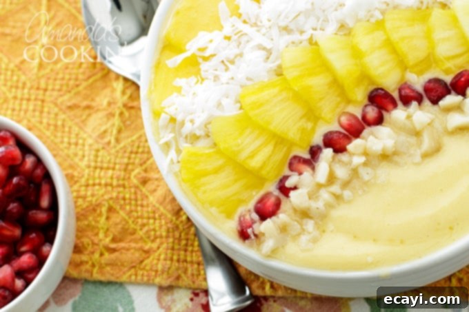 This refreshing tropical smoothie bowl is bursting with flavor and reminds me of a bowl of sunshine! This smoothie bowl is perfect for summer. #tropical #smoothie #smoothiebowl #breakfast #fruit #dairyfree #mango #peach #coconut Finished Tropical Smoothie Bowl, richly garnished with mango, pineapple, shredded coconut, pomegranate arils, and macadamia nuts
