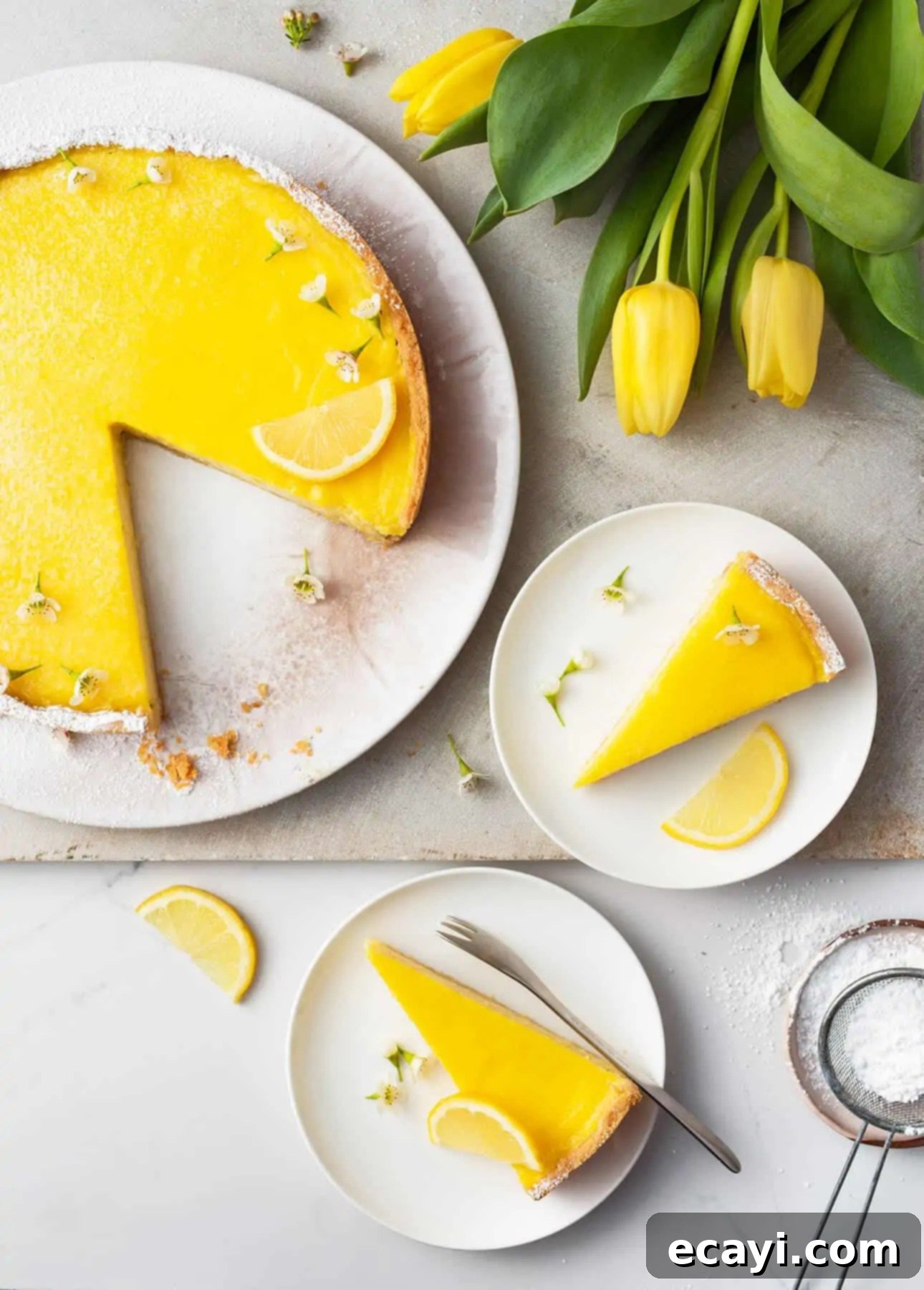 Authentic French Lemon Tart 2 A vibrant French lemon tart with a slice missing graces a round plate, accompanied by two smaller plates, each cradling a slice. Garnished with lemon slices and tiny flowers, its surrounded by sunny tulips and a sifter dusted in powdered sugar. // FoodNouveau.com