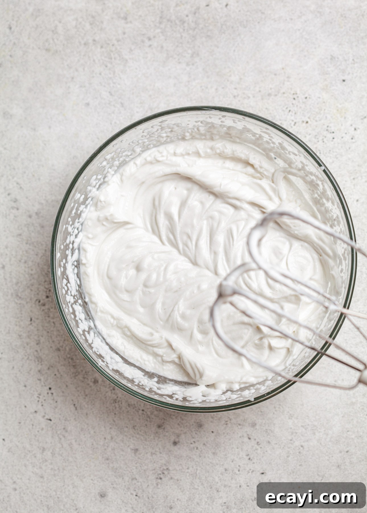 Authentic French Lemon Tart 8 A glass bowl filled with whipped Chantilly cream sits on a textured gray surface. An electric mixer whisk attachment rests on the edge, surrounded by fluffy, white cream. // FoodNouveau.com