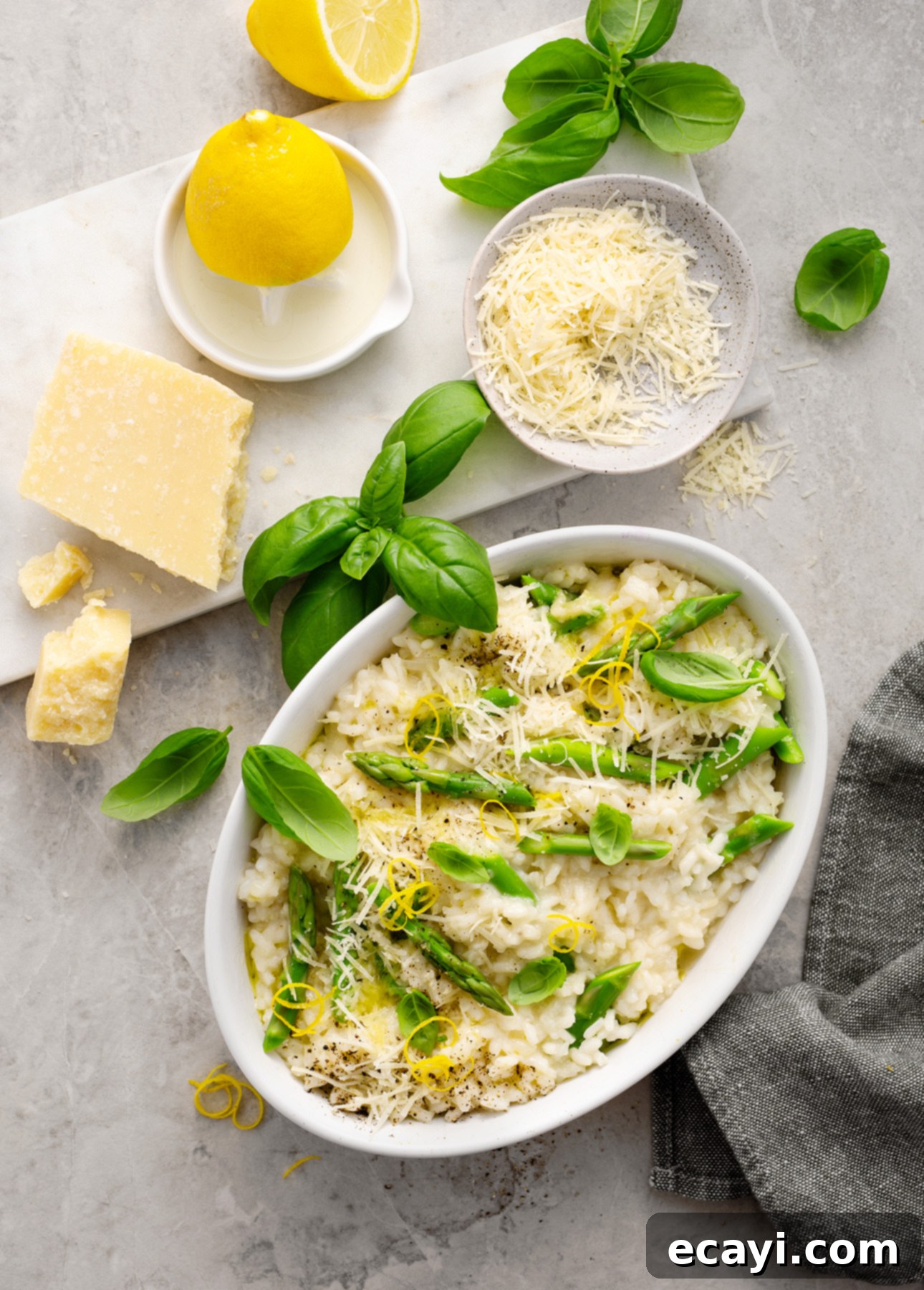 Creamy Lemon Asparagus Risotto 2 Oval dish of creamy lemon asparagus risotto garnished with lemon zest, grated cheese, and basil leaves. Surrounding the dish are a lemon, a cheese wedge, grated cheese, basil sprigs, and a gray napkin on a light surface. // FoodNouveau.com