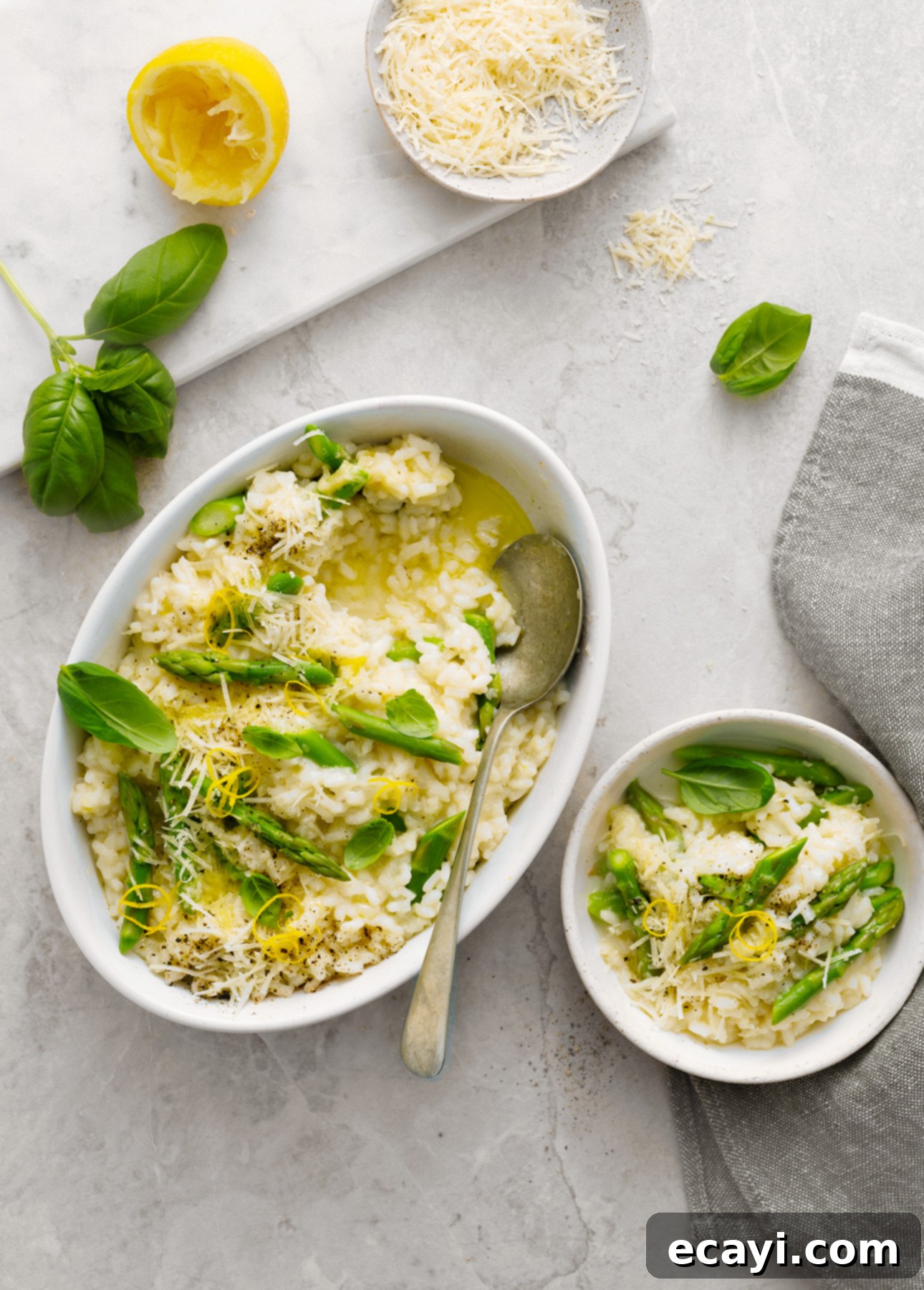 Creamy Lemon Asparagus Risotto 3 A dish and small bowl of creamy lemon asparagus risotto topped with basil leaves and grated cheese sit on a light surface. Nearby are a piece of lemon, fresh basil, extra grated cheese, and a gray napkin. A spoon rests in the main dish. // FoodNouveau.com