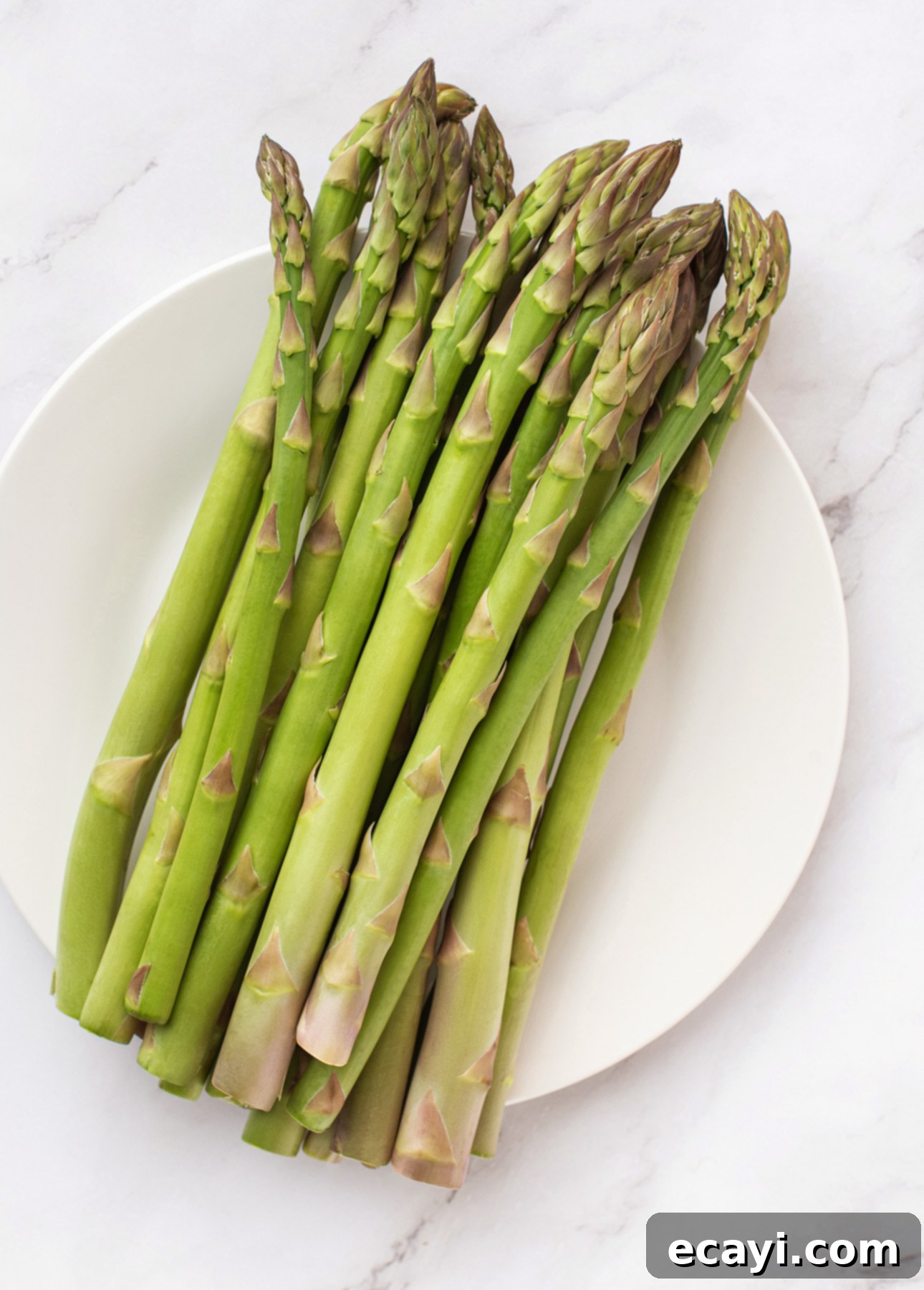 Creamy Lemon Asparagus Risotto 7 A bundle of fresh green asparagus spears arranged on a white plate, set on a light marble surface—perfect for preparing a vibrant lemon asparagus risotto. // FoodNouveau.com