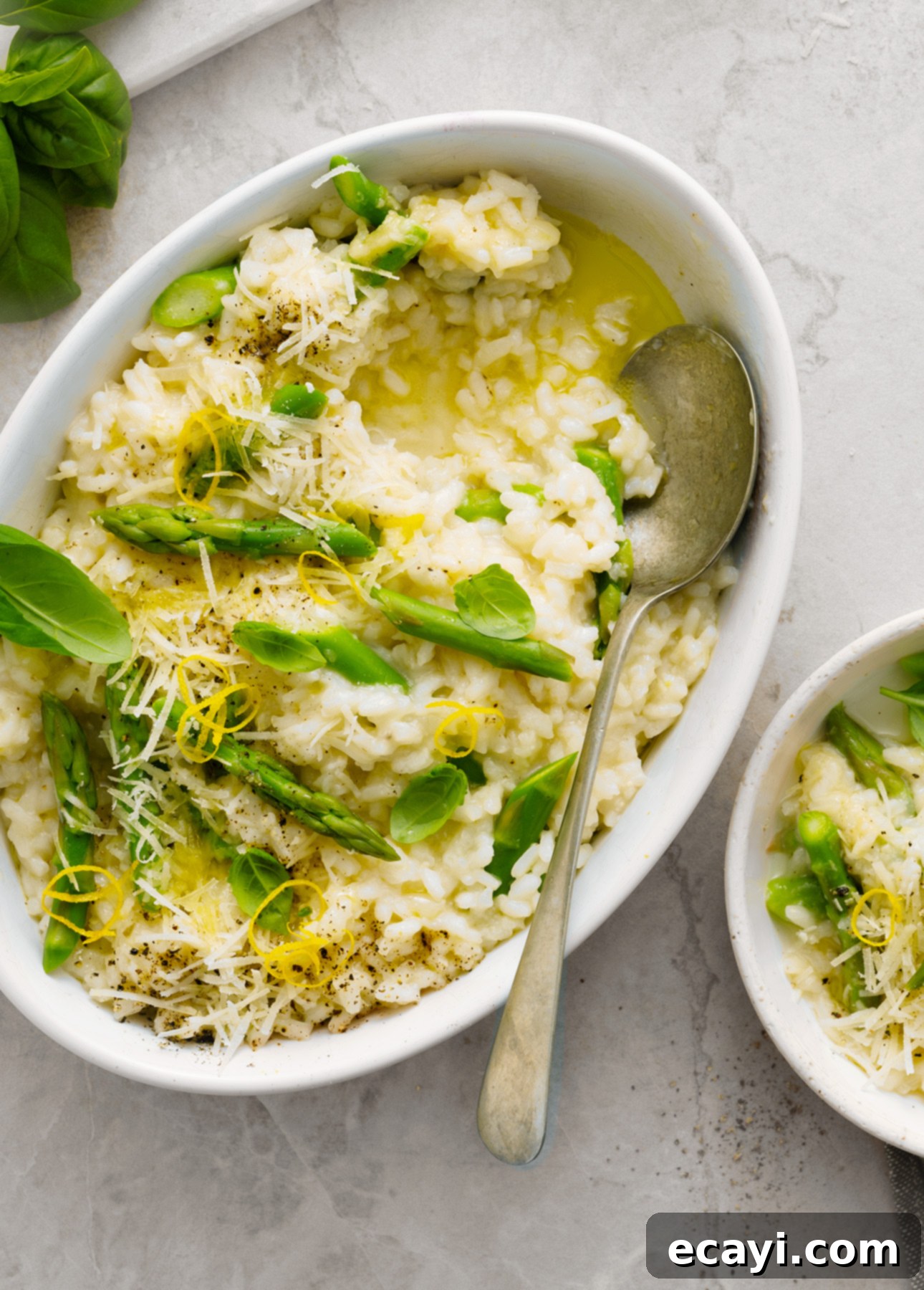 Creamy Lemon Asparagus Risotto 8 Oval dish of creamy lemon asparagus risotto garnished with grated cheese, fresh basil, lemon zest, black pepper, and a drizzle of olive oil, with a spoon resting inside. A small serving sits on a plate nearby. // FoodNouveau.com