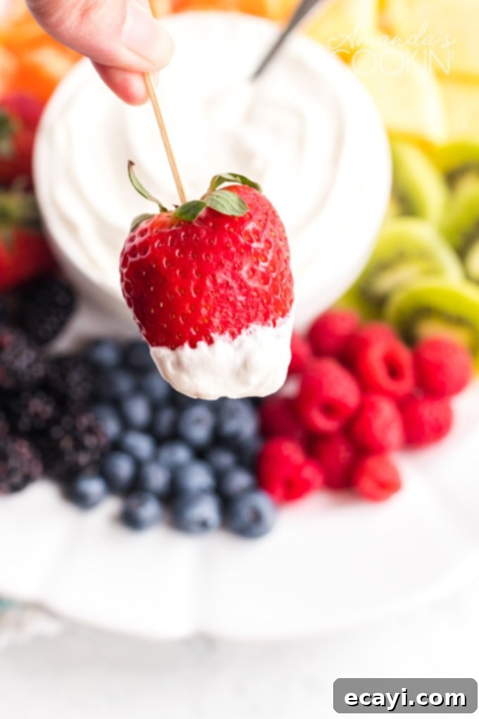This Cream Cheese Fruit Dip is a really cool way to sneak in lots of fresh fruit on hot summer days. It's sweet and fluffy- perfect with any summer fruit! A hand holding a vibrant red strawberry on a toothpick, dipped generously into the creamy white fruit dip, against a blurred background of more fruit.