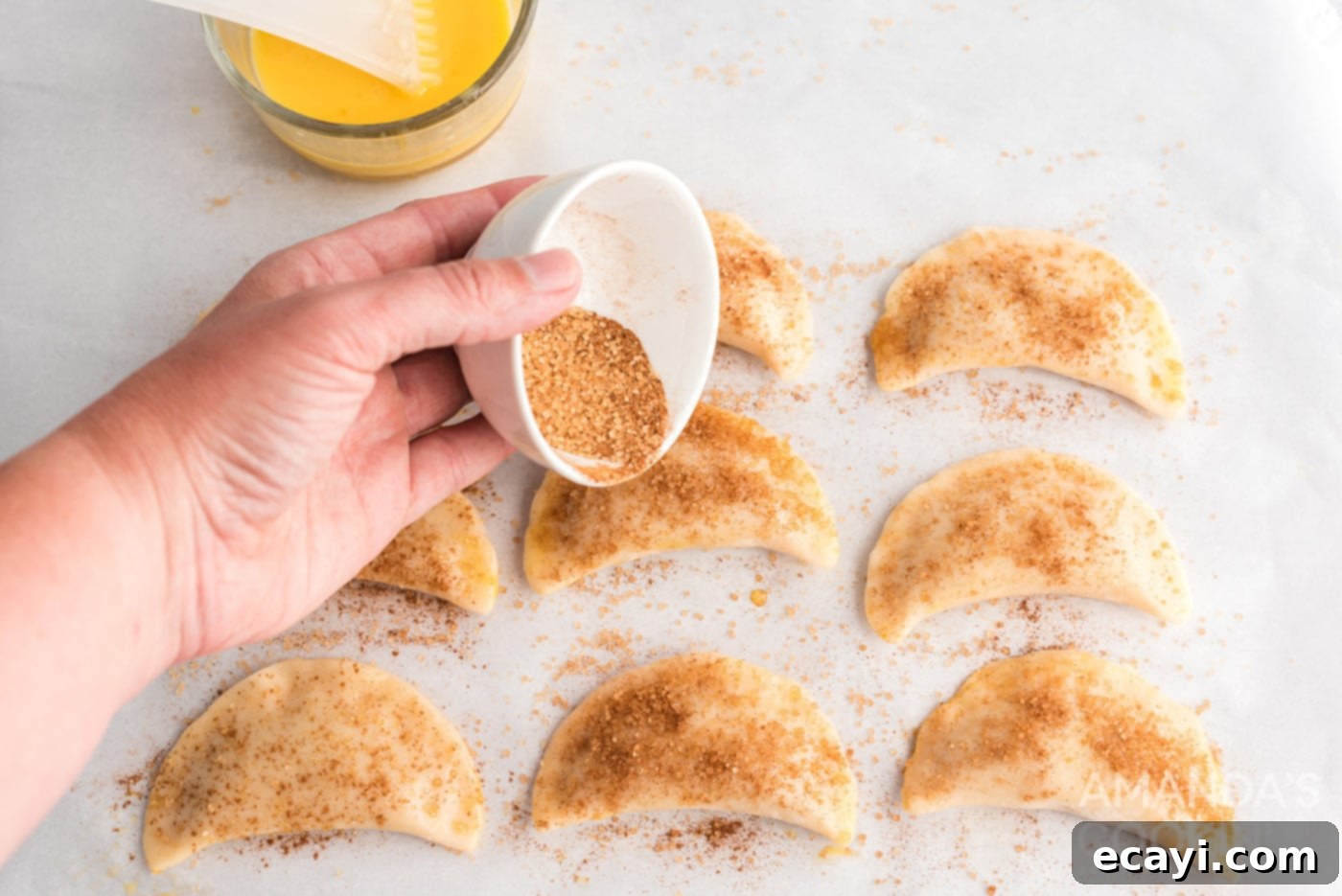 Flaky Pumpkin Turnovers 10 Cinnamon sugar being sprinkled generously over pumpkin hand pies arranged on a baking sheet, ready for the oven.
