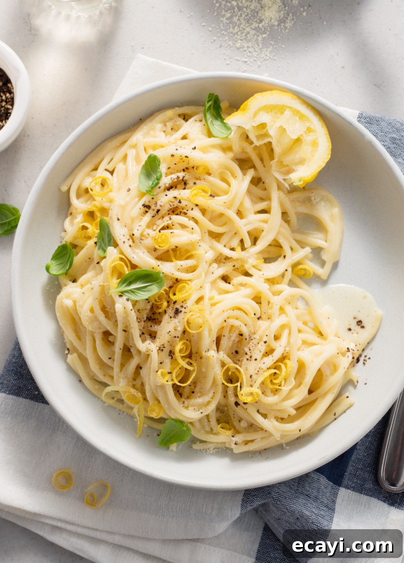 Zesty Lemon Cream Spaghetti 7 A inviting bowl of creamy spaghetti al limone is presented, garnished with vibrant fresh basil leaves, a dusting of freshly cracked black pepper, aromatic lemon zest, and a bright lemon wedge. It's served on a pristine white plate, which rests atop a charming blue and white striped napkin. // FoodNouveau.com