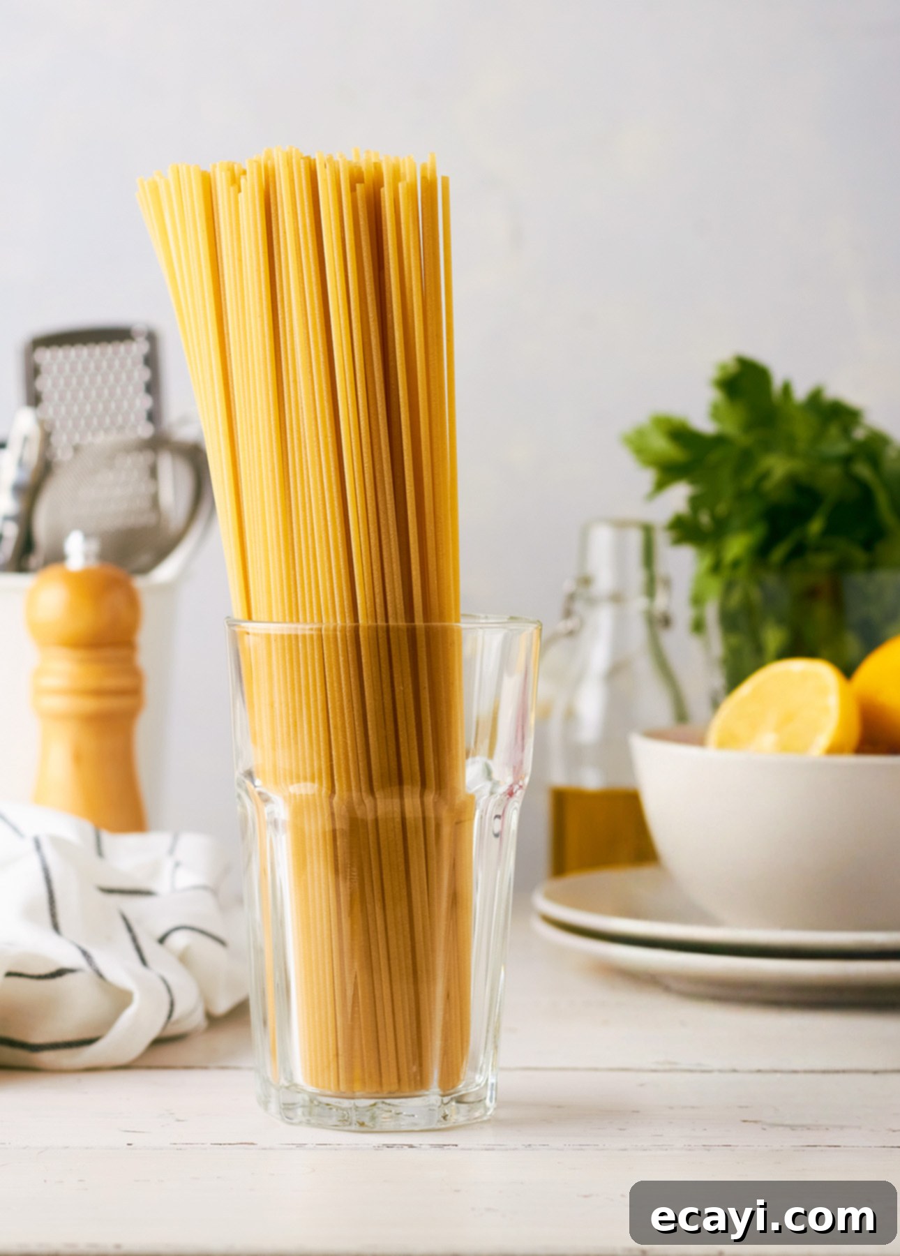 Zesty Lemon Cream Spaghetti 8 A tall glass filled with uncooked spaghetti strands stands proudly on a bright white kitchen table, surrounded by an inviting array of fresh ingredients. A striped cloth, a robust pepper grinder, a trusty cheese grater, vibrant fresh parsley, and a whole lemon are perfectly arranged – all essential components for crafting the most delectable creamy lemon pasta. A pristine white bowl and an elegant bottle of olive oil complete the scene, hinting at the culinary magic about to unfold. // FoodNouveau.com