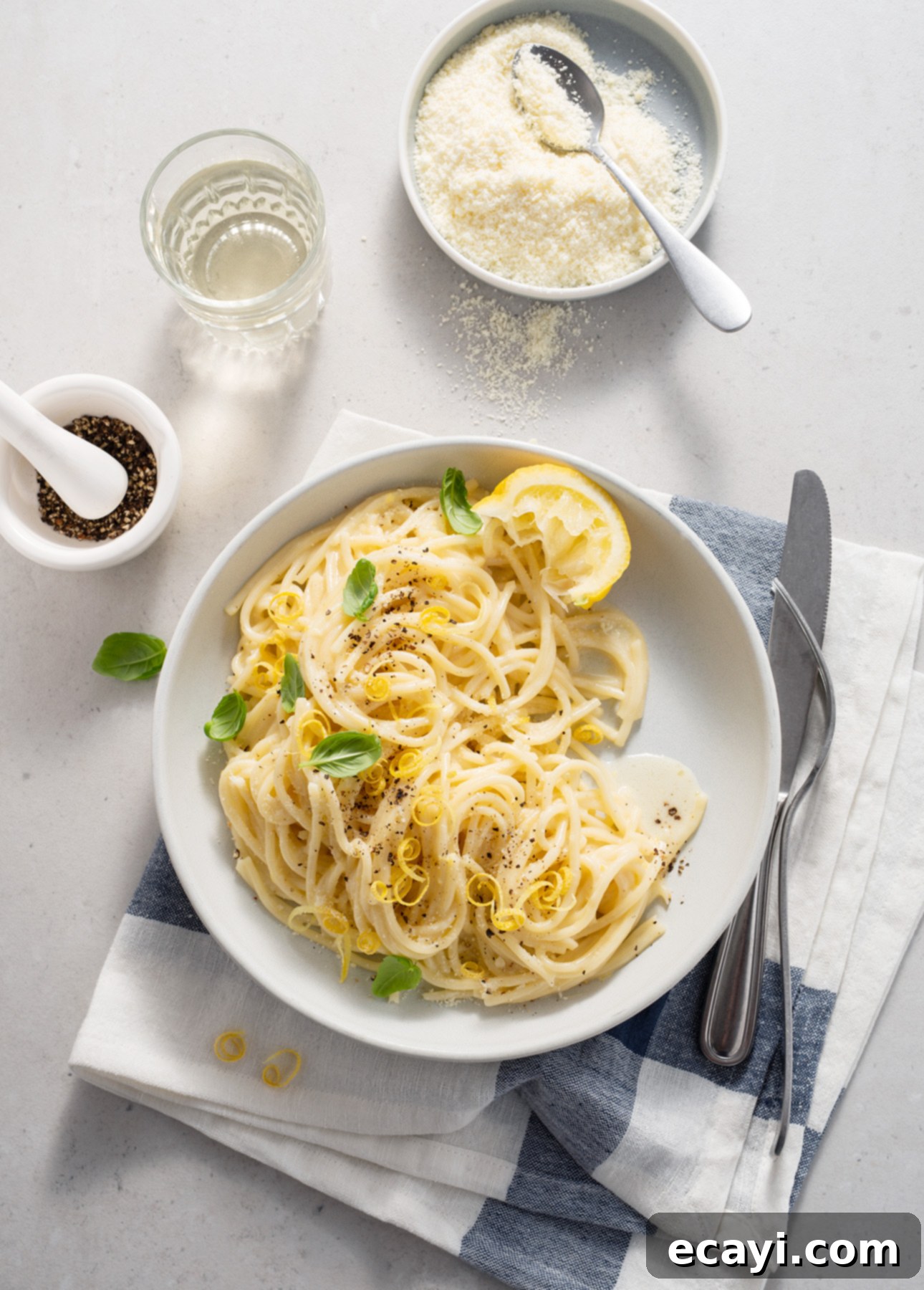 Zesty Lemon Cream Spaghetti 9 A delectable plate of creamy spaghetti al limone, elegantly garnished with vibrant lemon slices, fresh basil leaves, and a sprinkle of black pepper. The dish is presented on a clean white napkin, accompanied by a fork and knife. Nearby, a small bowl of grated cheese, a refreshing glass of water, and a mortar with whole peppercorns complete the inviting culinary scene. // FoodNouveau.com