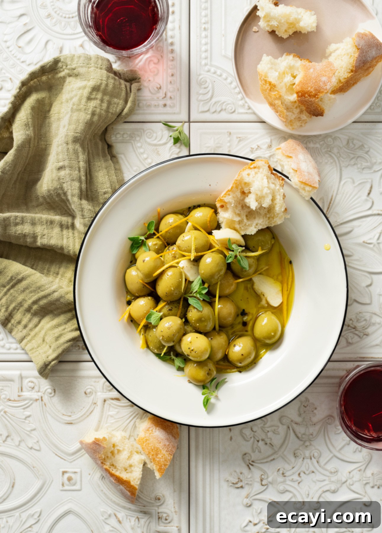 Zesty Warm Olives 3 A bowl of warm citrus olives, garnished with fresh herbs and citrus peel, accompanied by a small dish of the flavorful marinade, set on a rustic wooden surface.