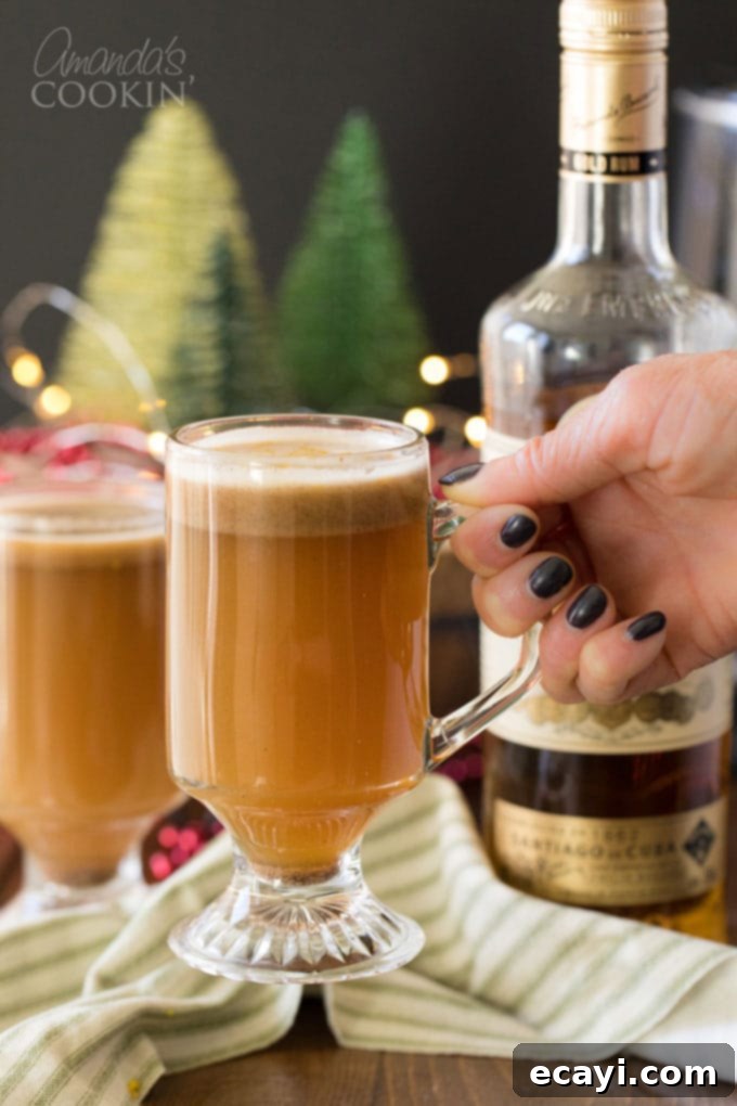 A Hot Buttered Rum - AKA a hot toddy - is a dessert-like cocktail with a rich, buttery flavor that is gently spiced and finished off with a twist of orange. woman's hand picking up glass of hot buttered rum
