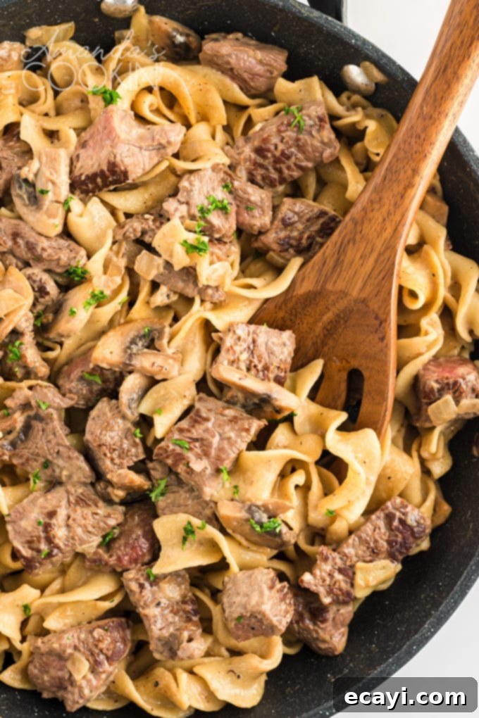 This classic beef stroganoff recipe from scratch features tender slices of steak and savory mushrooms smothered in creamy brown gravy that's ready in 30! #beefrecipes #beefstroganoff #pasta #pastarecipes #dinnerrecipes #mushrooms #skillet #skilletdinners #easydinnerrecipes #noodles pan of beef stroganoff with wooden spoon