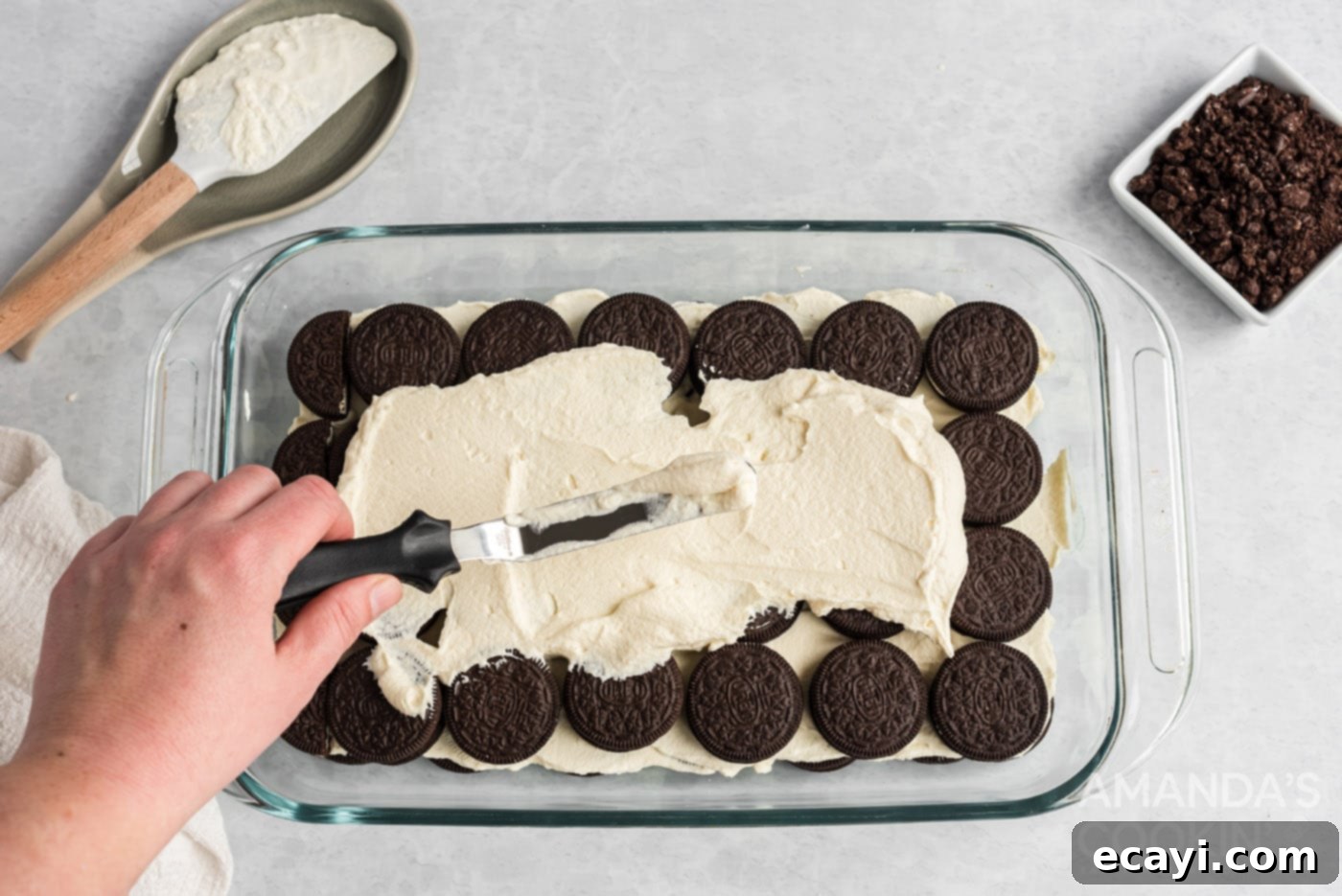 No-Bake Oreo Delight 7 Smooth, homemade whipped cream being carefully spread over the first layer of Oreo cookies in a baking dish.