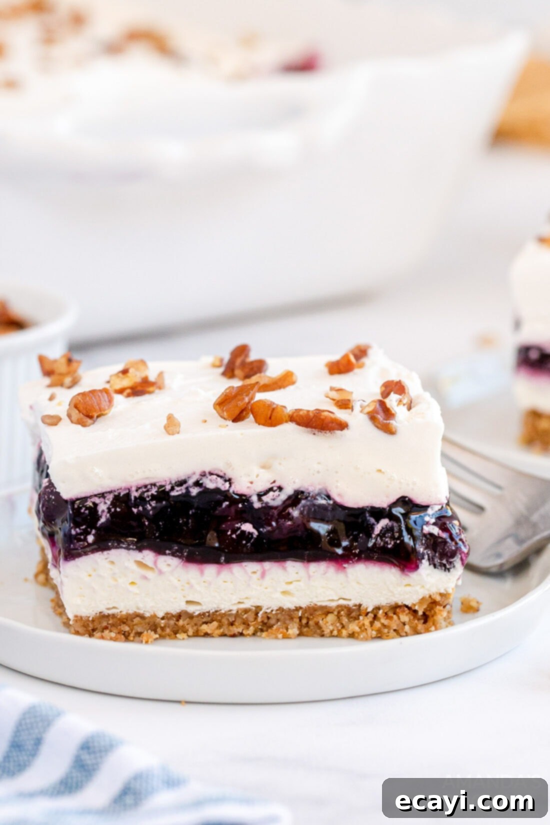 Blueberry Bliss 14 A perfectly cut slice of blueberry lush on a white plate, showcasing its distinct layers of crust, cream cheese, blueberry, and whipped topping.