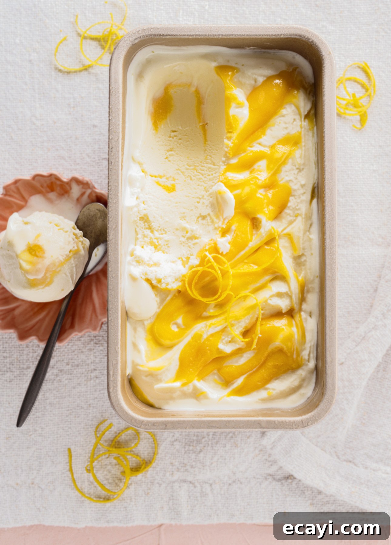 Zesty Homemade Lemon Gelato 11 A rectangular dish of gelato swirled with bright yellow lemon curd is placed on a textured white cloth. Next to it, a coral-colored dish holds a scoop of gelato. Lemon zest curls are artfully scattered around, enhancing the citrus allure. // FoodNouveau.com