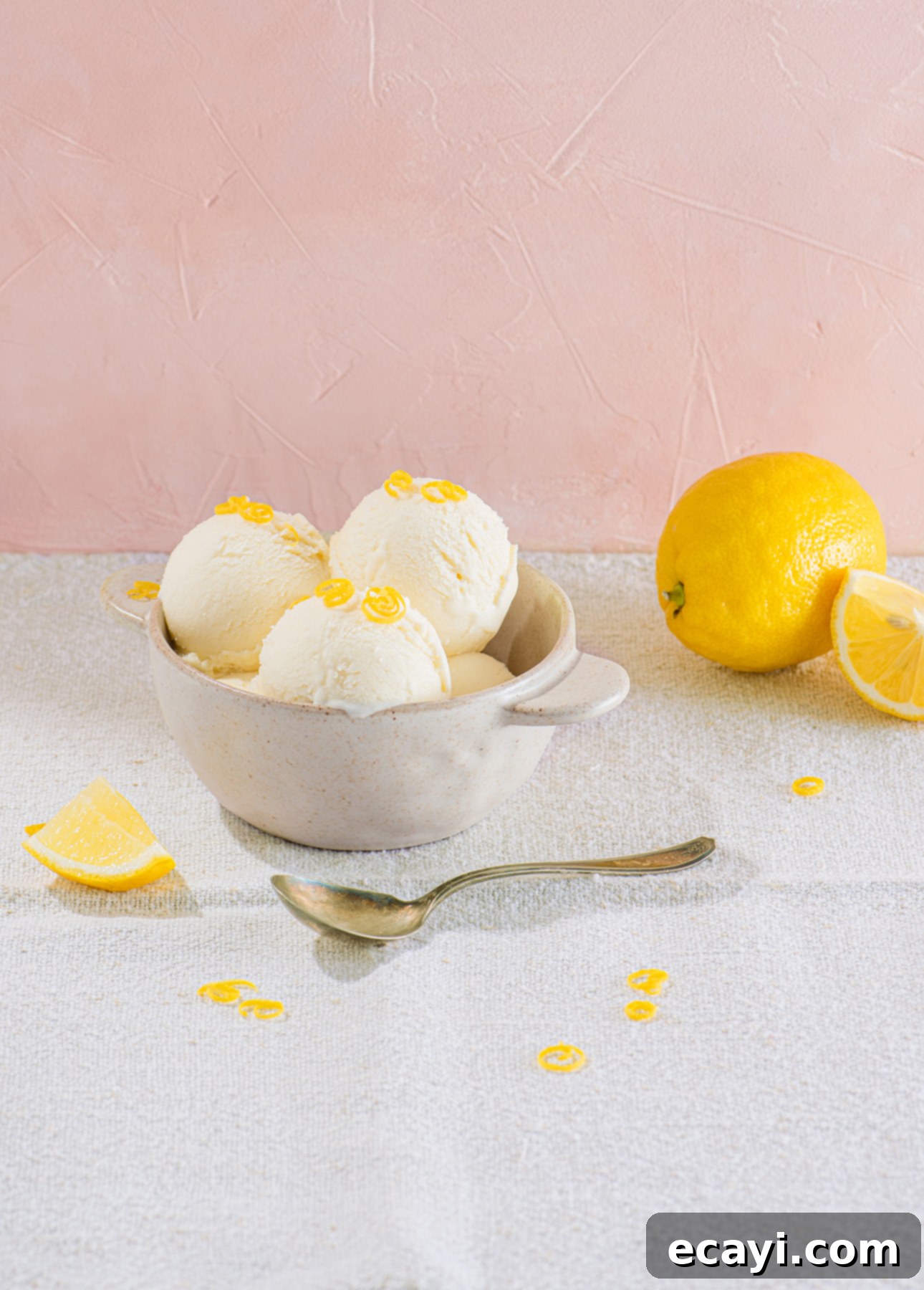 Zesty Homemade Lemon Gelato 12 A ceramic bowl with three scoops of gelato topped with lemon zest rests on textured light fabric. Nearby, a whole lemon and a lemon wedge add zesty flair. A spoon lies in front of the bowl against the soft pink background. // FoodNouveau.com