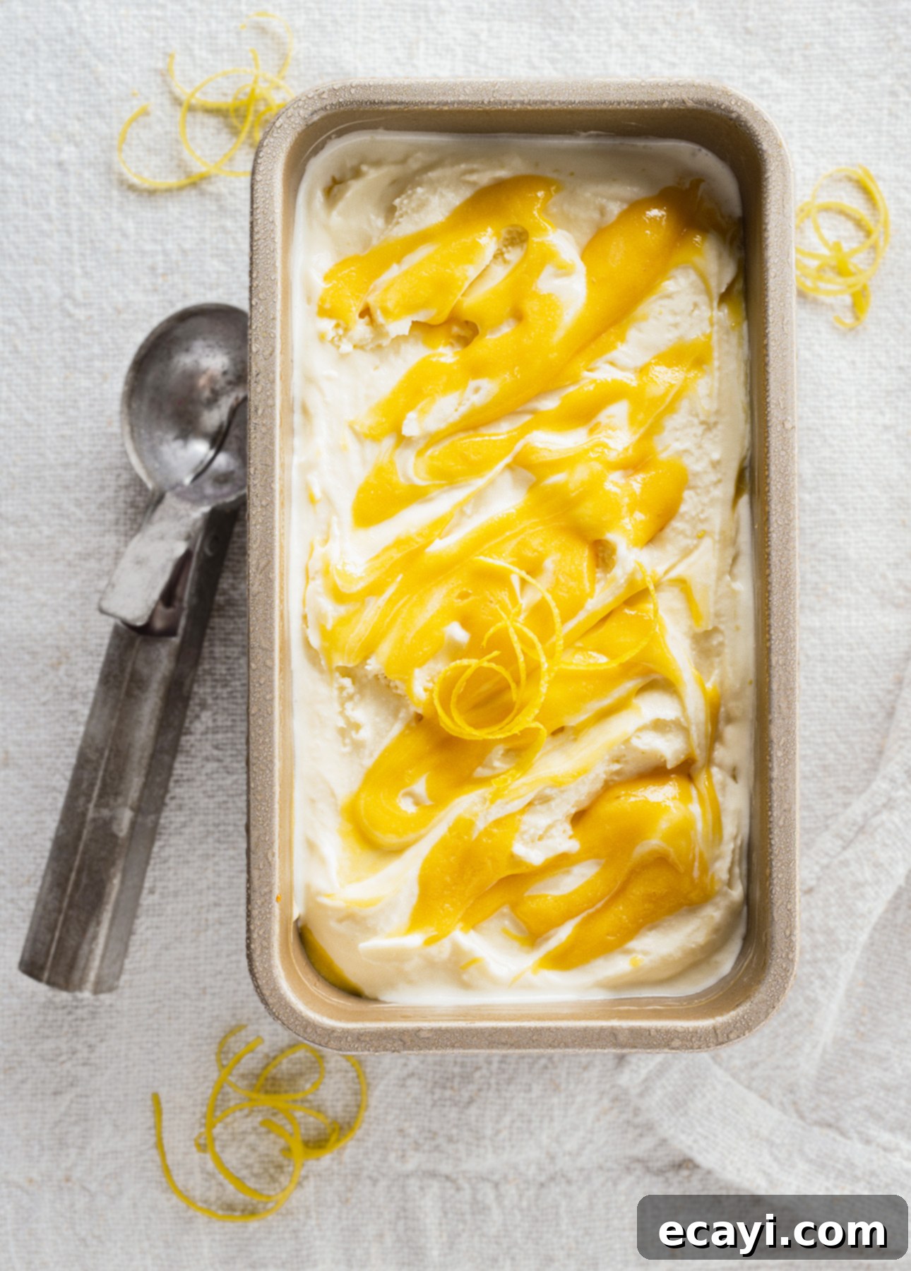 Zesty Homemade Lemon Gelato 5 A tin container filled with creamy gelato swirled with vibrant yellow lemon curd. A metal ice cream scoop rests beside the container on a textured white cloth, with lemon zest sprinkled decoratively around. // FoodNouveau.com