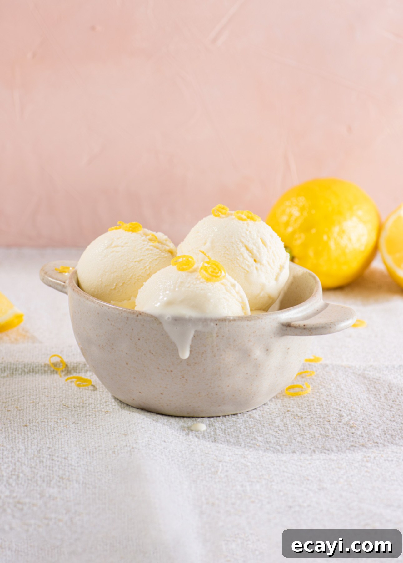 Zesty Homemade Lemon Gelato 6 A ceramic bowl with three scoops of creamy gelato garnished with lemon zest. Two whole lemons sit in the background on a light textured surface, while a soft pink wall adds warmth. A bit of gelato drips over the edge of the bowl. // FoodNouveau.com