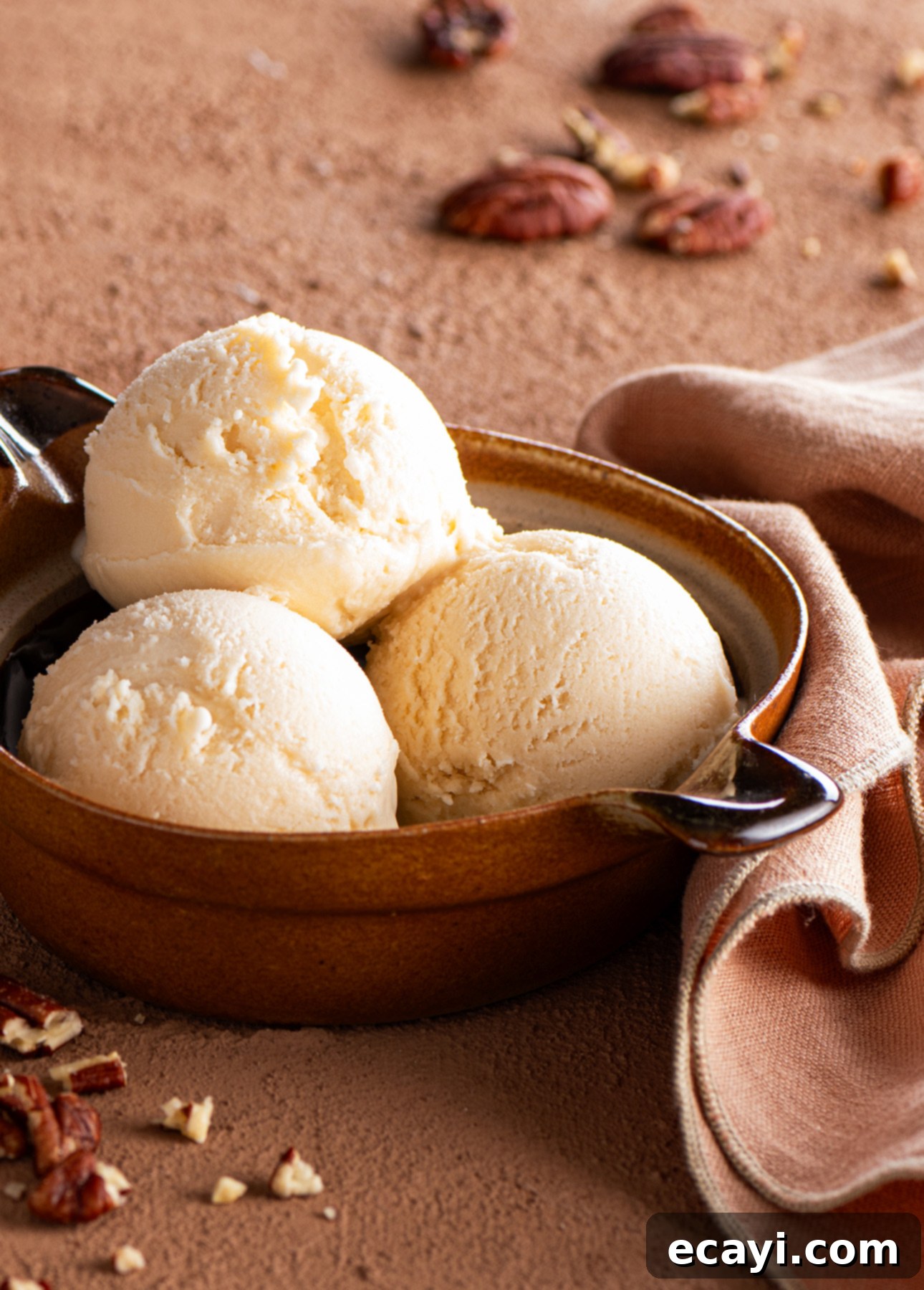 Silky Maple Bliss 3 A brown bowl holds three scoops of creamy maple gelato, tempting you with its rich sweetness. On a brown surface scattered with chopped pecans, the bowl invites indulgence. A peach-colored cloth draped nearby enhances the warmth. // FoodNouveau.com