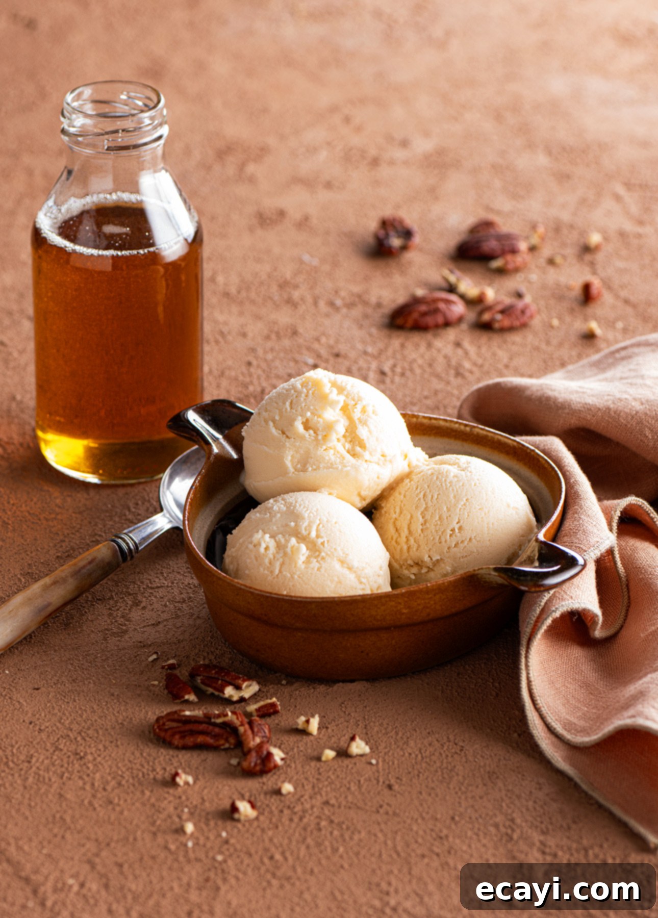 Silky Maple Bliss 5 A bowl with three scoops of maple gelato sits on a brown surface, surrounded by scattered pecans. A glass bottle of syrup and a beige napkin with a spoon rest nearby. // FoodNouveau.com