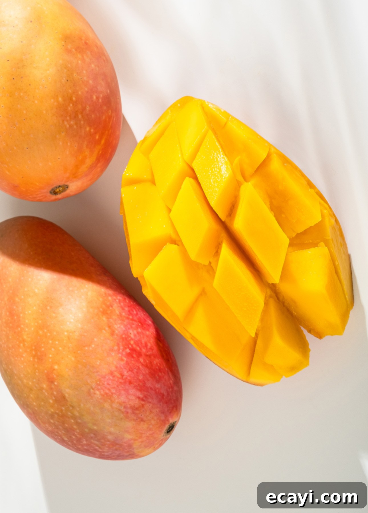 Tropical Vegan Passion Fruit Mango Gelato 4 Two whole mangoes and one halved and cubed mango are displayed on a white surface. The mango on the left is partially in shadow, while the cubed mango reveals vibrant yellow flesh. // FoodNouveau.com