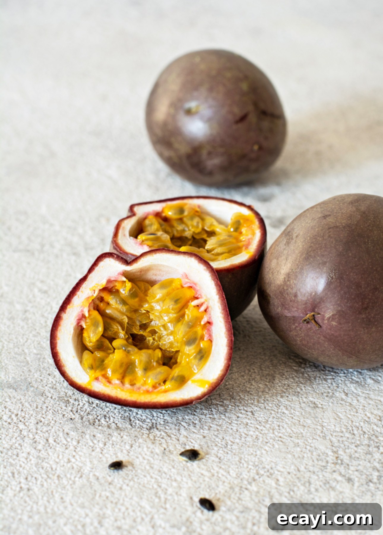 Tropical Vegan Passion Fruit Mango Gelato 5 Two whole passion fruits and one halved fruit rest on a light gray surface, revealing juicy, yellow pulp dotted with seeds. A few seeds are scattered about. // FoodNouveau.com