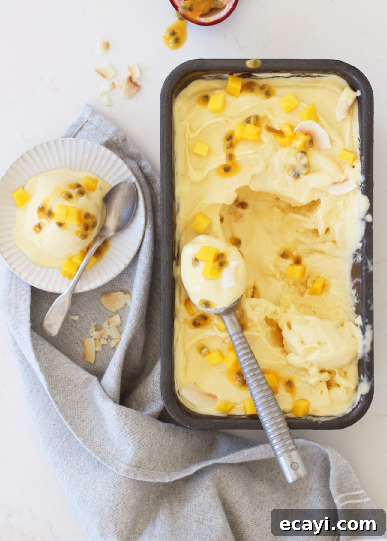 Tropical Vegan Passion Fruit Mango Gelato 6 A tub of mango gelato, decorated with passion fruit and mango chunks, is partially scooped into a bowl nearby. A gray cloth is artistically draped on the marble surface, with spoons and coconut flakes scattered around. // FoodNouveau.com