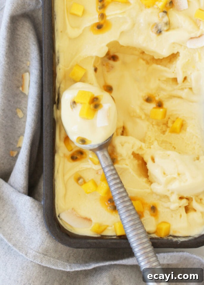 Tropical Vegan Passion Fruit Mango Gelato 8 A close-up of creamy yellow mango gelato adorned with chunks of mango and passion fruit seeds, elegantly served in a metal container. A spoon rests on the edge, capturing a perfect scoop against a light gray cloth background. // FoodNouveau.com