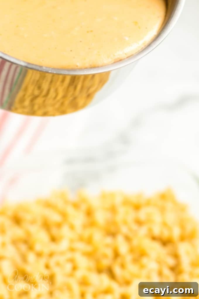 Homemade baked macaroni and cheese is pure comfort food, just like grandma used to make. This baked mac and cheese recipe is easy to make and uses a delicious homemade creamy cheese sauce and is topped with crunchy bread crumbs. I'll show you how to make good old fashioned baked macaroni and cheese that's a true classic! #bakedmacaroniandcheese #bakedmacandcheese #macaroniandcheese #easymacandcheese #homemademacandcheese #howtomakemacaroniandcheese #elbowmacaroni #cheddarcheese #casserolerecipe Step-by-step image showing creamy cheese sauce being poured over cooked elbow macaroni in a baking dish, ready for the next stage of assembly.