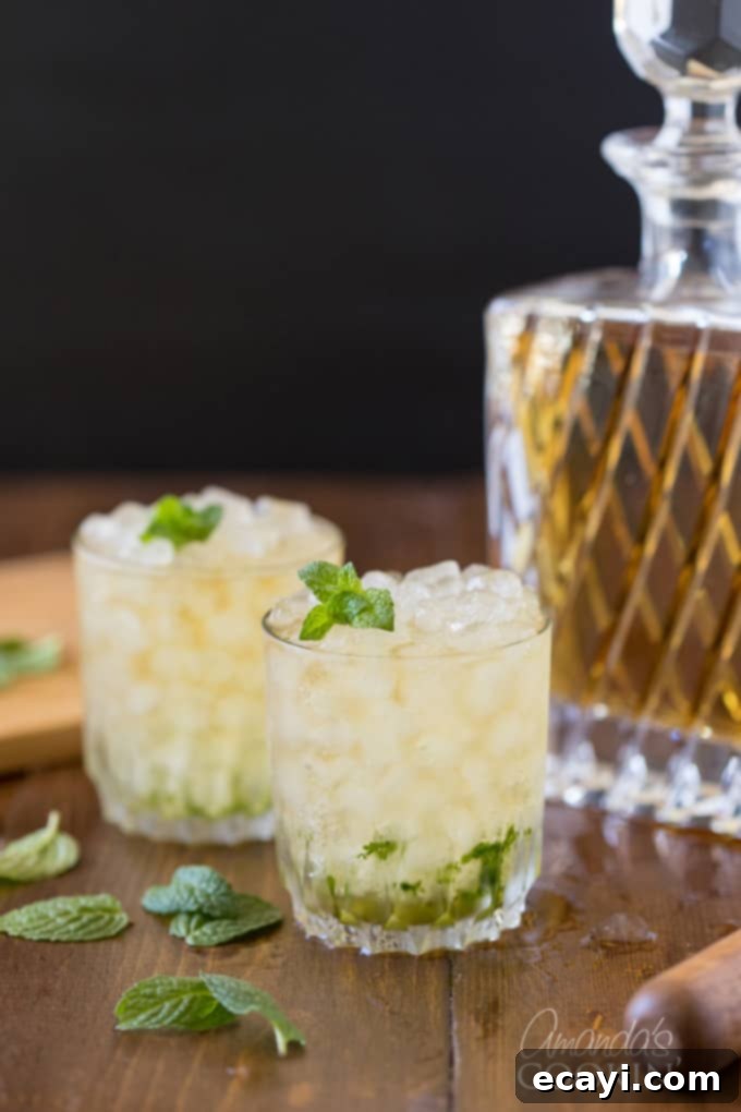 The classic Mint Julep, signature drink of the Kentucky Derby, is made with bourbon whiskey and served in a highball glass or a silver julep cup. Discover this easy cocktail recipe for an authentic taste of Kentucky. Mint Julep with whiskey bottle in background, highlighting the key ingredients of bourbon and mint.