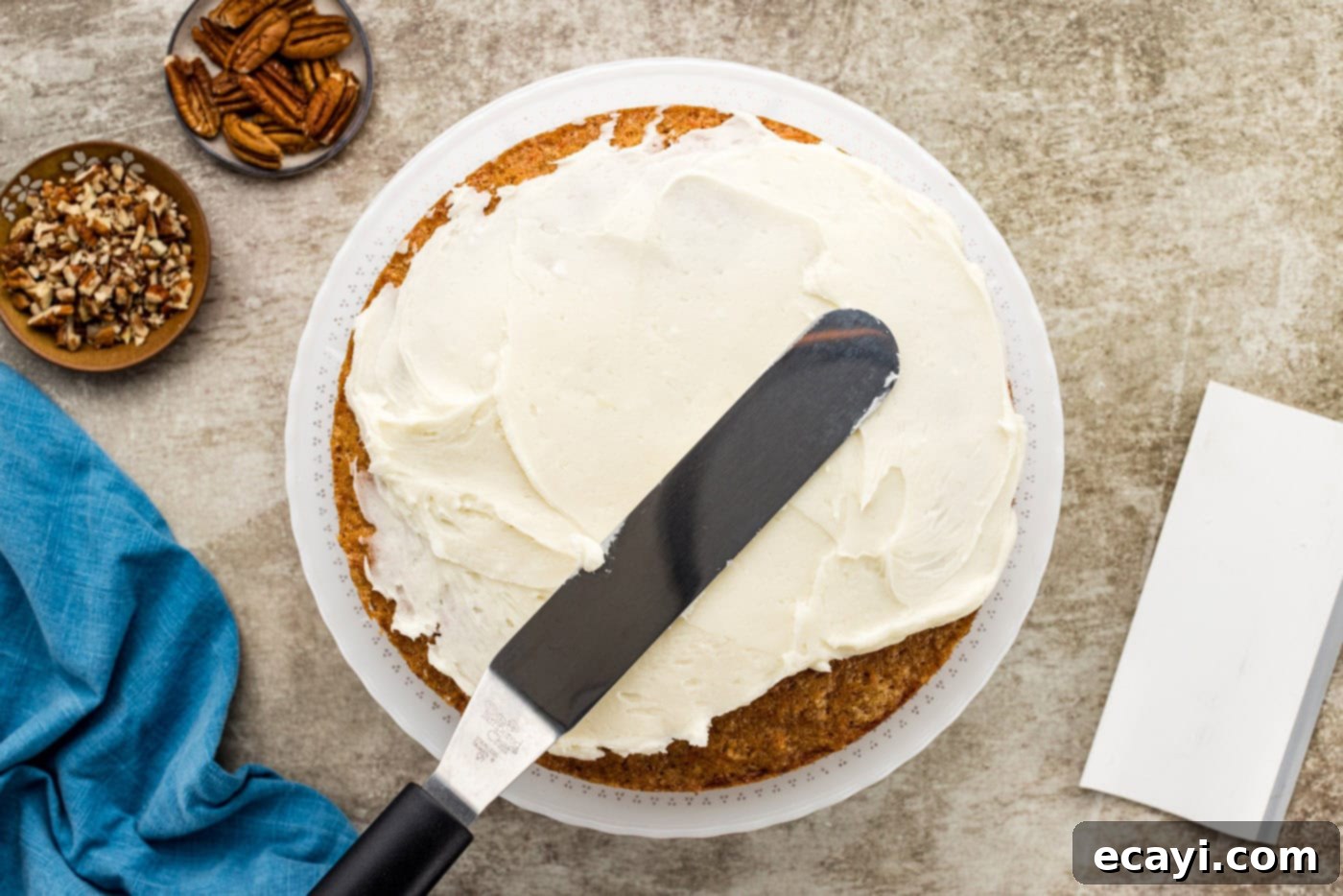 Spiced Carrot Delight 10 An icing spatula smoothly spreading creamy white frosting over the sides of a stacked carrot cake, creating a crumb coat.