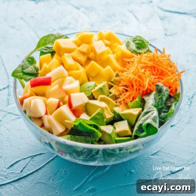 This delicious spinach salad is packed with protein-rich leafy spinach, sweet mango, and healthy fats from avocado. Perfect for summer! #spinachsalad #avocadosalad #mangosalad #summersalad #leafygreens #freshsaladrecipes #spinachsaladrecipes #meatlesssalad #amandascookin bowl of fresh salad ingredients including spinach, carrots, mango, avocado, and apple, ready for preparation