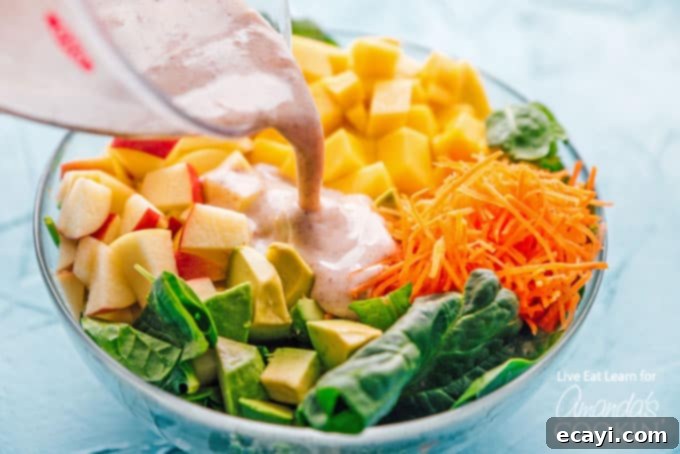 This delicious spinach salad is packed with protein-rich leafy spinach, sweet mango, and healthy fats from avocado. Perfect for summer! #spinachsalad #avocadosalad #mangosalad #summersalad #leafygreens #freshsaladrecipes #spinachsaladrecipes #meatlesssalad #amandascookin Pouring the creamy Greek yogurt dressing over a fresh spinach salad with mango, avocado, and apple