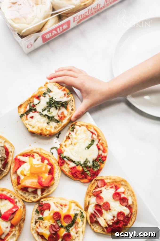 Muffin Pizza Creation Bar 2 child's hand reaching for a freshly baked English muffin pizza, showcasing golden cheese and toppings