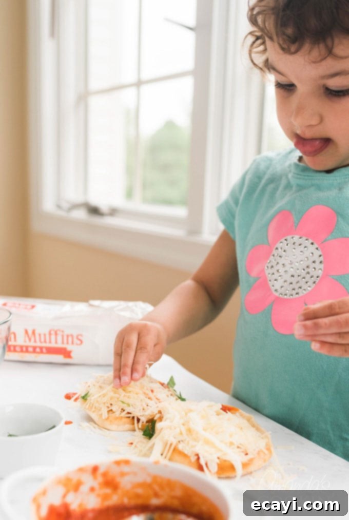Muffin Pizza Creation Bar 7 Small child diligently adding shredded cheese to an English muffin pizza, demonstrating intense concentration