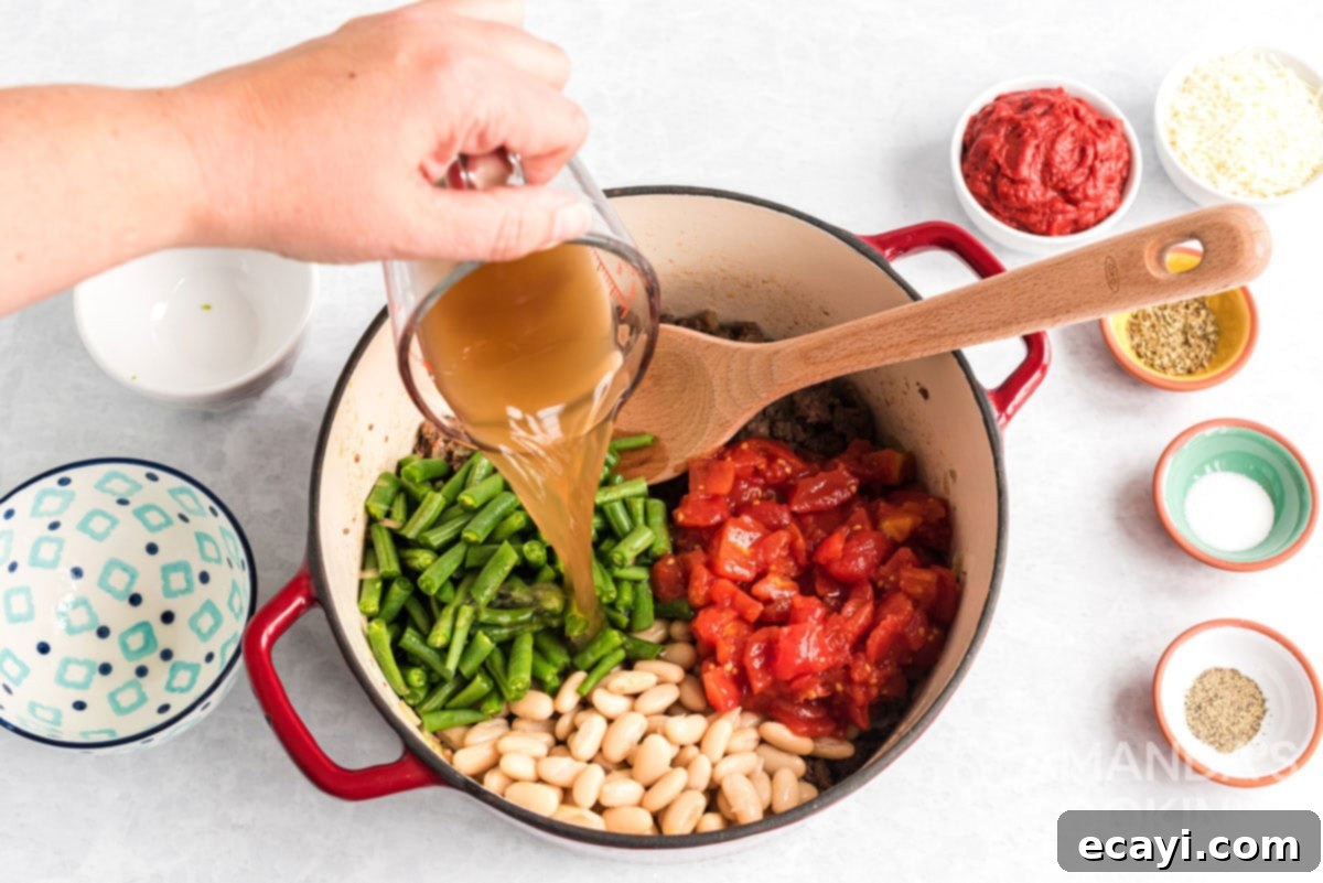 Hearty Ground Beef & Vegetable Stew 4 Pouring vegetable broth into a Dutch oven filled with browned ground meat, onions, beans, and tomatoes for Italian hamburger stew, blending the flavors.