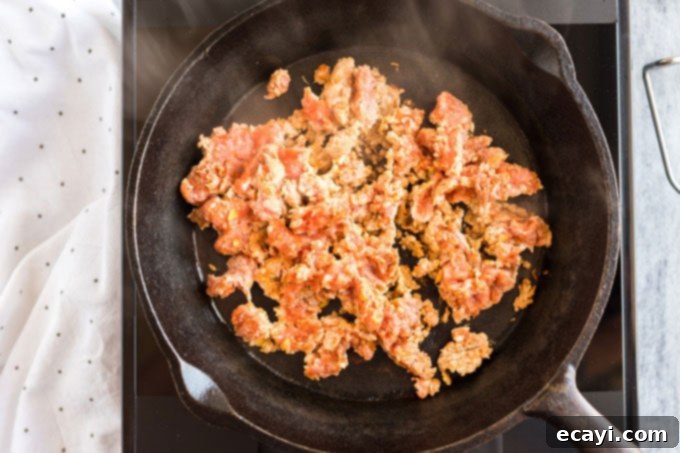 Crafting Authentic Pizza Sausage 7 Ground pizza sausage being browned in a hot cast iron skillet, with a wooden spoon breaking up the meat for even cooking.