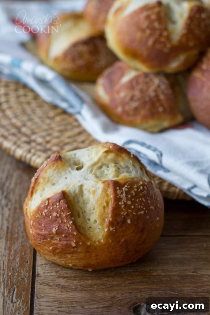Pretzel Rolls 2 A close up of a bretzel roll with other rolls laying on a white and blue towel in the background, highlighting its soft texture and golden-brown crust.