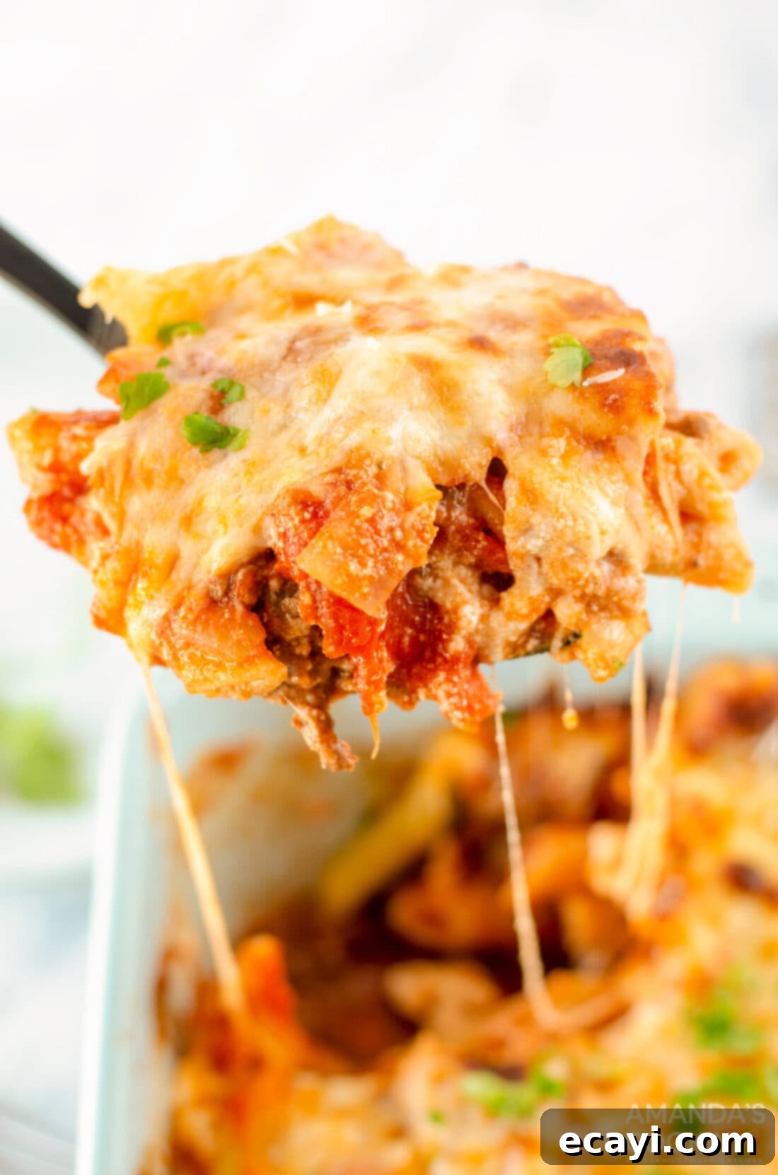 Ultimate Cheesy Ziti Bake 3 A serving spoon lifting a portion of baked ziti, showing layers of pasta, meat sauce, and cheese