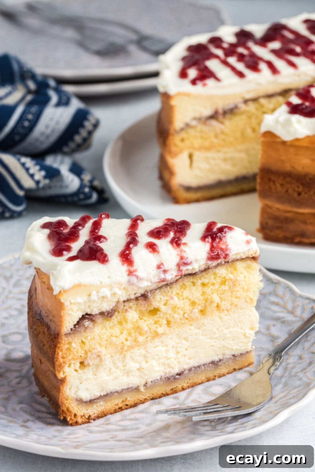 Regal Trifle Cheesecake 2 A beautiful slice of English Trifle Cheesecake showing off its distinct layers of yellow cake, raspberry, and creamy cheesecake, topped with whipped cream and a raspberry drizzle.