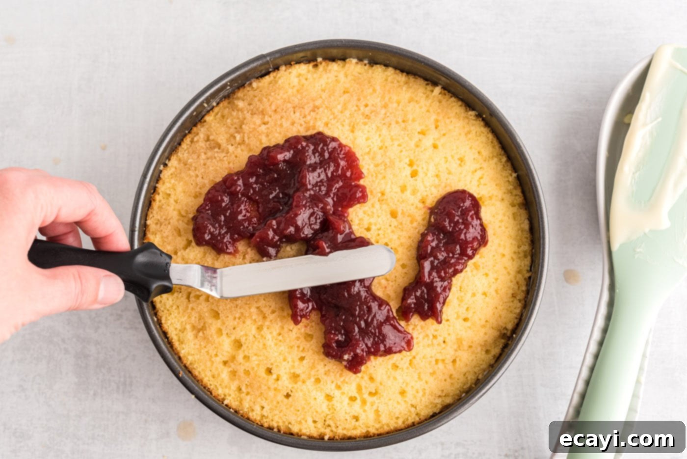 Regal Trifle Cheesecake 10 An icing spatula spreading raspberry preserves smoothly over the second layer of yellow cake in the springform pan.