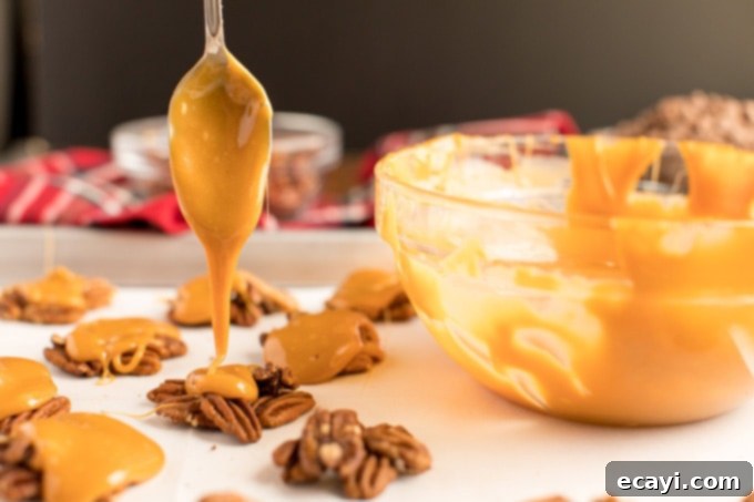 Applying gooey caramel to pecan clusters for the homemade chocolate turtle recipe. A spoon drizzling melted caramel over pecan mounds on a baking sheet