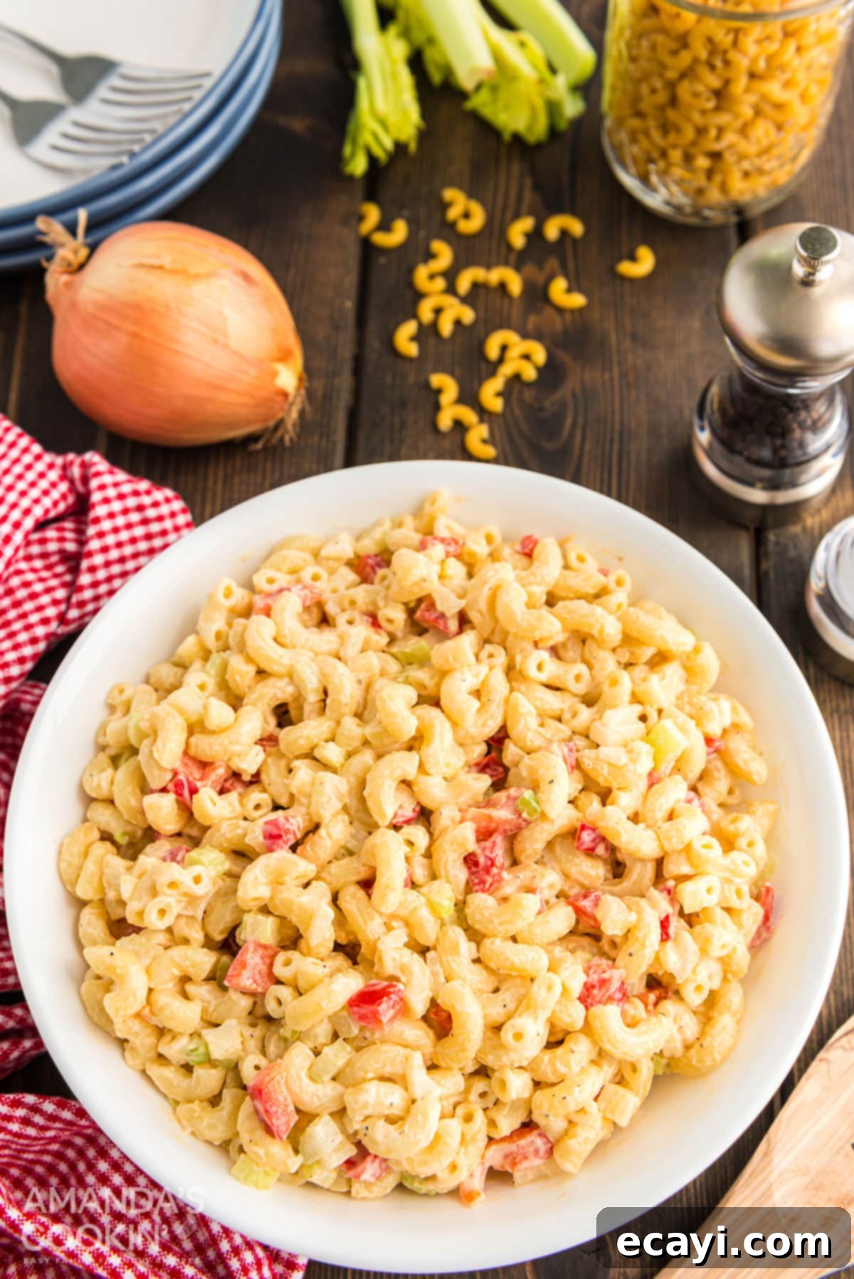 A close-up shot of the finished macaroni salad, with fresh ingredients in the background. A bowl of creamy macaroni salad is placed on a rustic wooden table, with whole peppercorns, onion, and dried macaroni pasta in the soft-focus background, suggesting the ingredients.